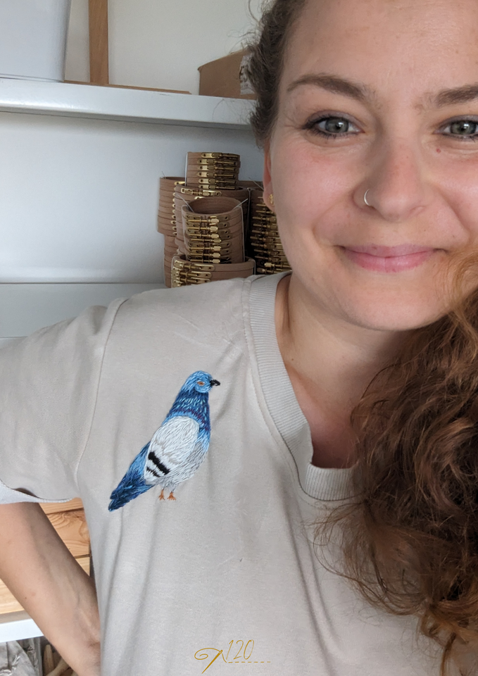 Gründerin von "Reparieren ist Liebe" Vera Teske lächelnd mit einem gestickten Vogel auf der Schulter ihres T-shirts